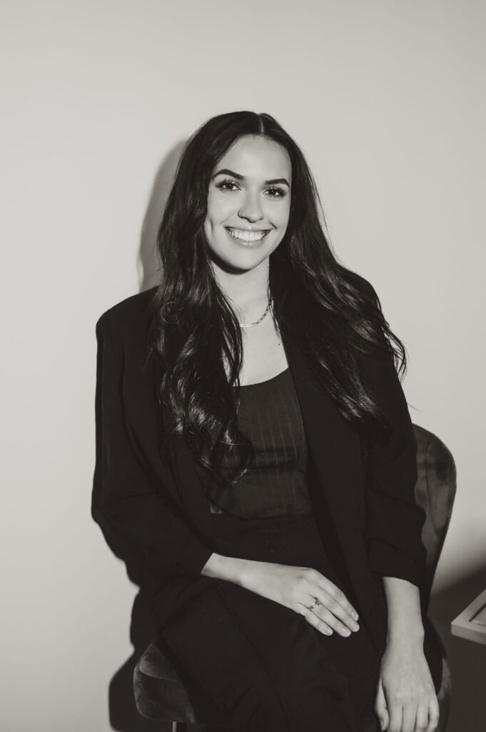 Smiling person sitting, wearing black blazer and pants
